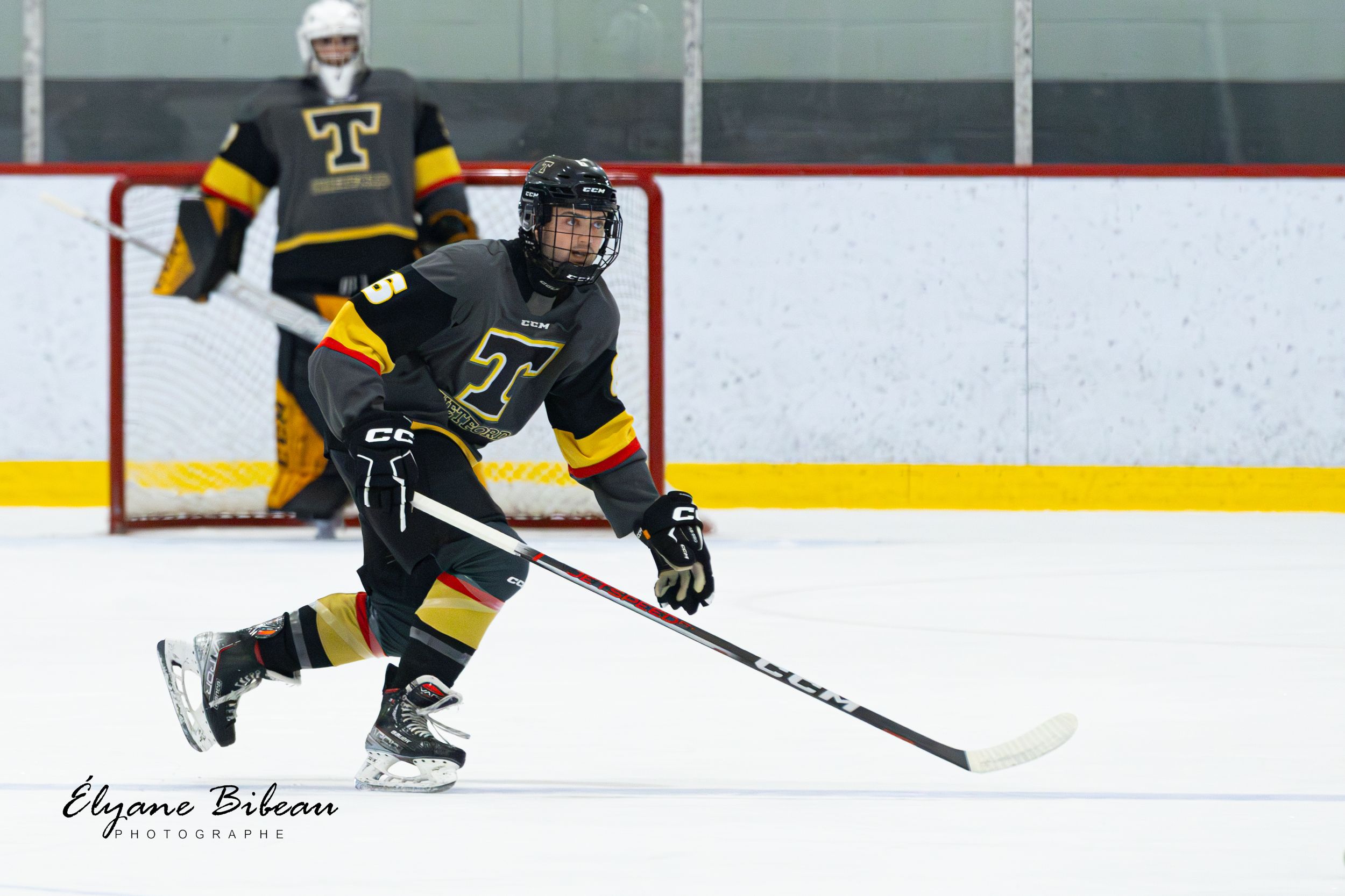 Hockey player in black and gold uniform carries puck across ice with teammate in goal behind.