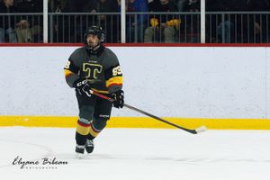 Hockey player in dark uniform with yellow accents skates during game action.