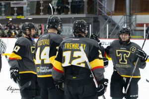 Hockey team huddles on ice wearing matching black and yellow uniforms with numbers.