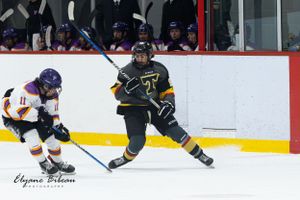 Ice hockey player in black and yellow jersey skates with the puck while opponent in purple pursues.