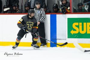 Hockey player in black uniform battles for puck against boards while referee watches.