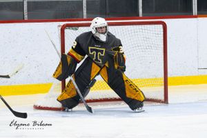 Goalie in black and gold uniform crouches in ready position at ice hockey net.