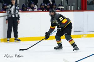 Hockey player in black uniform handles puck while skating on ice with referee in background.