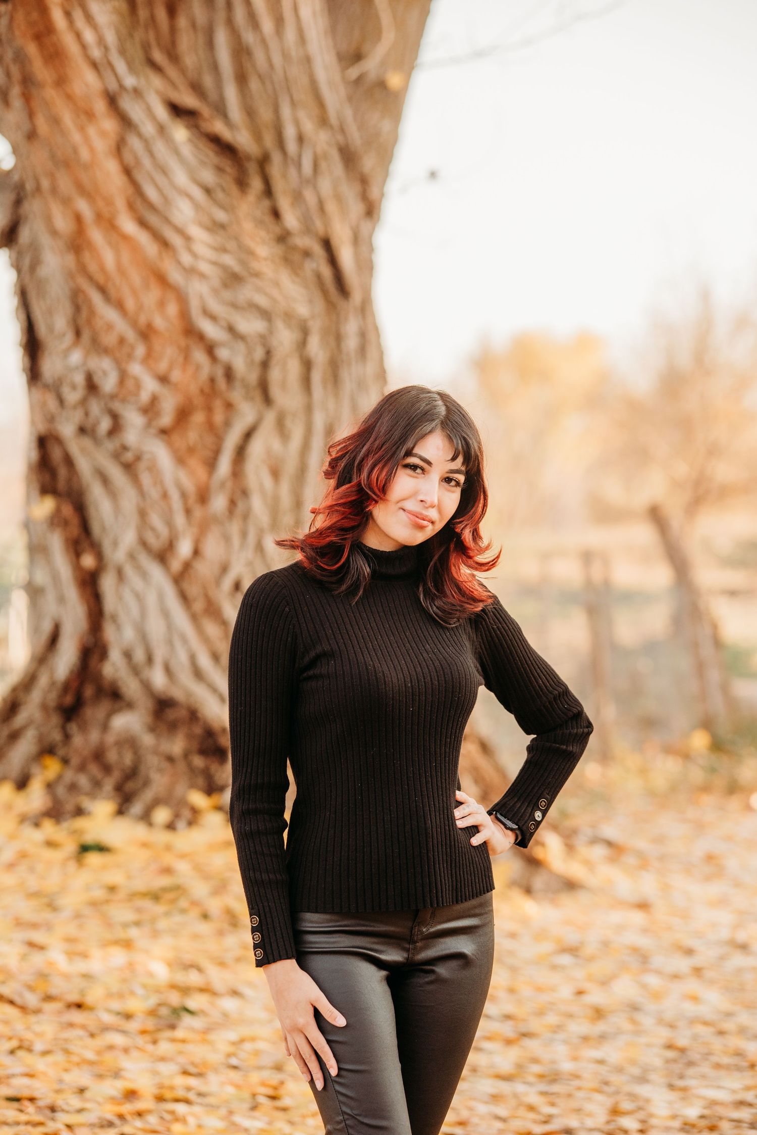 Woman in black sweater poses against textured tree trunk with autumn foliage in background.