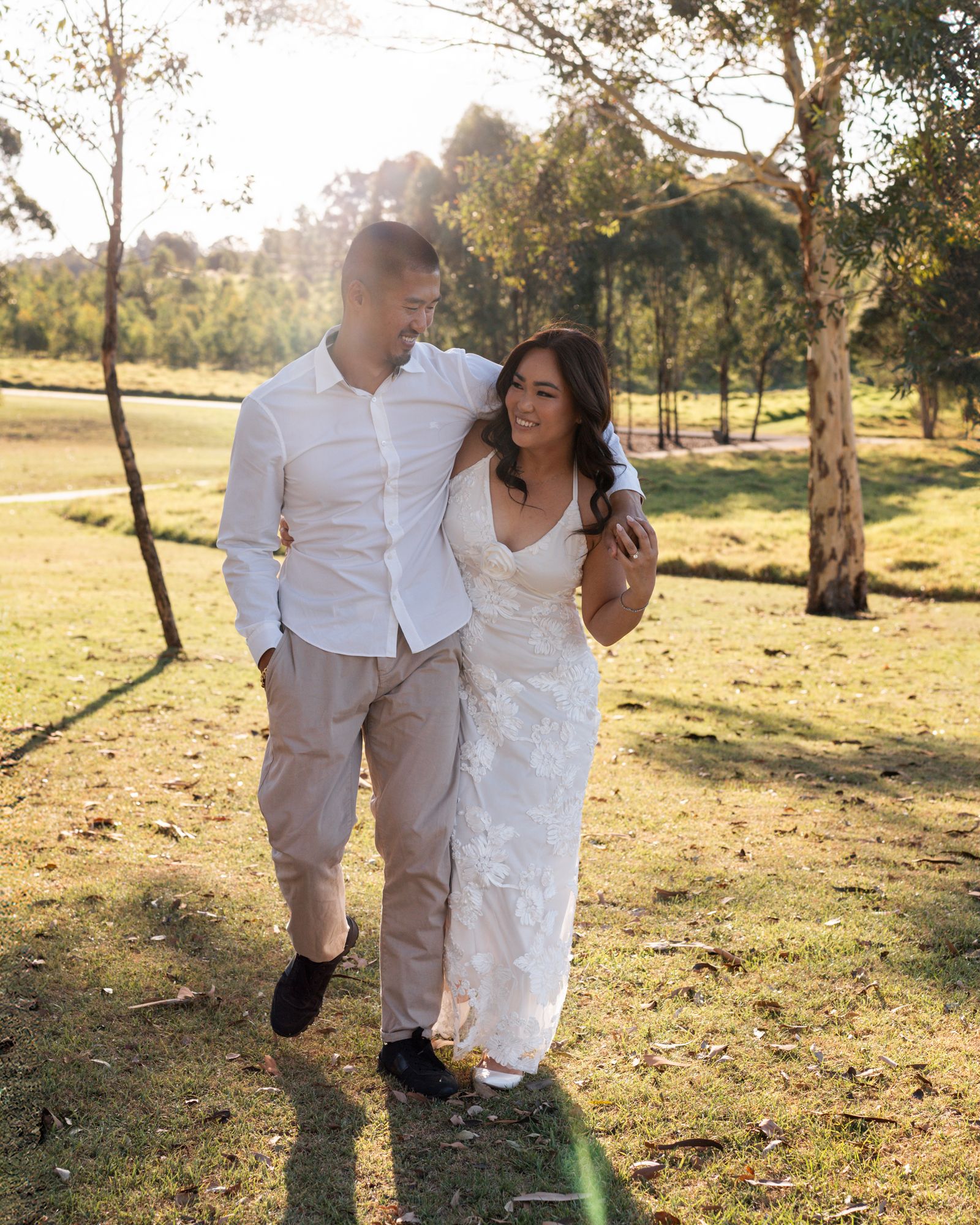 A couple in white outfits playfully walks through a sunlit outdoor field during a romantic photoshoot.