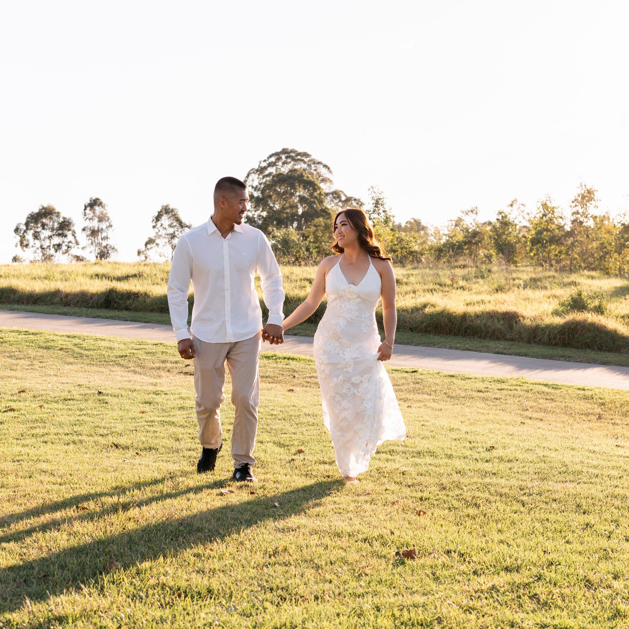 A couple strolls hand in hand across a sunlit grassy field during a sunset photoshoot.