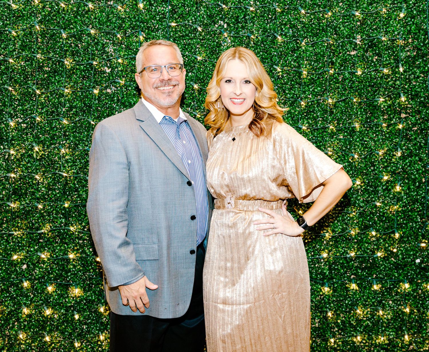 Two people pose together against a green hedge wall backdrop at an event, wearing formal attire.