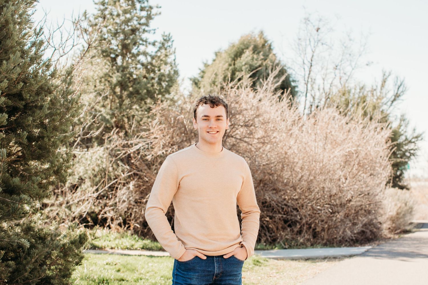 Young person in beige sweater standing outdoors on sunny day with bushes in background.
