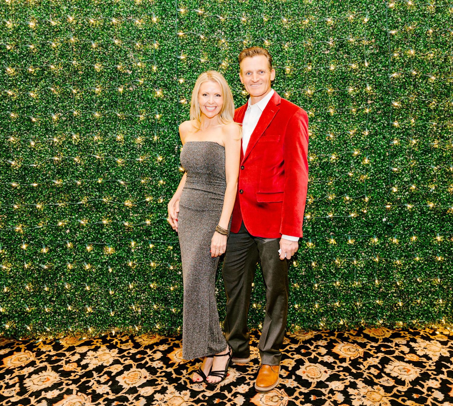Two people pose together against a green floral backdrop, with one wearing a red blazer and the other in a sparkly gown.