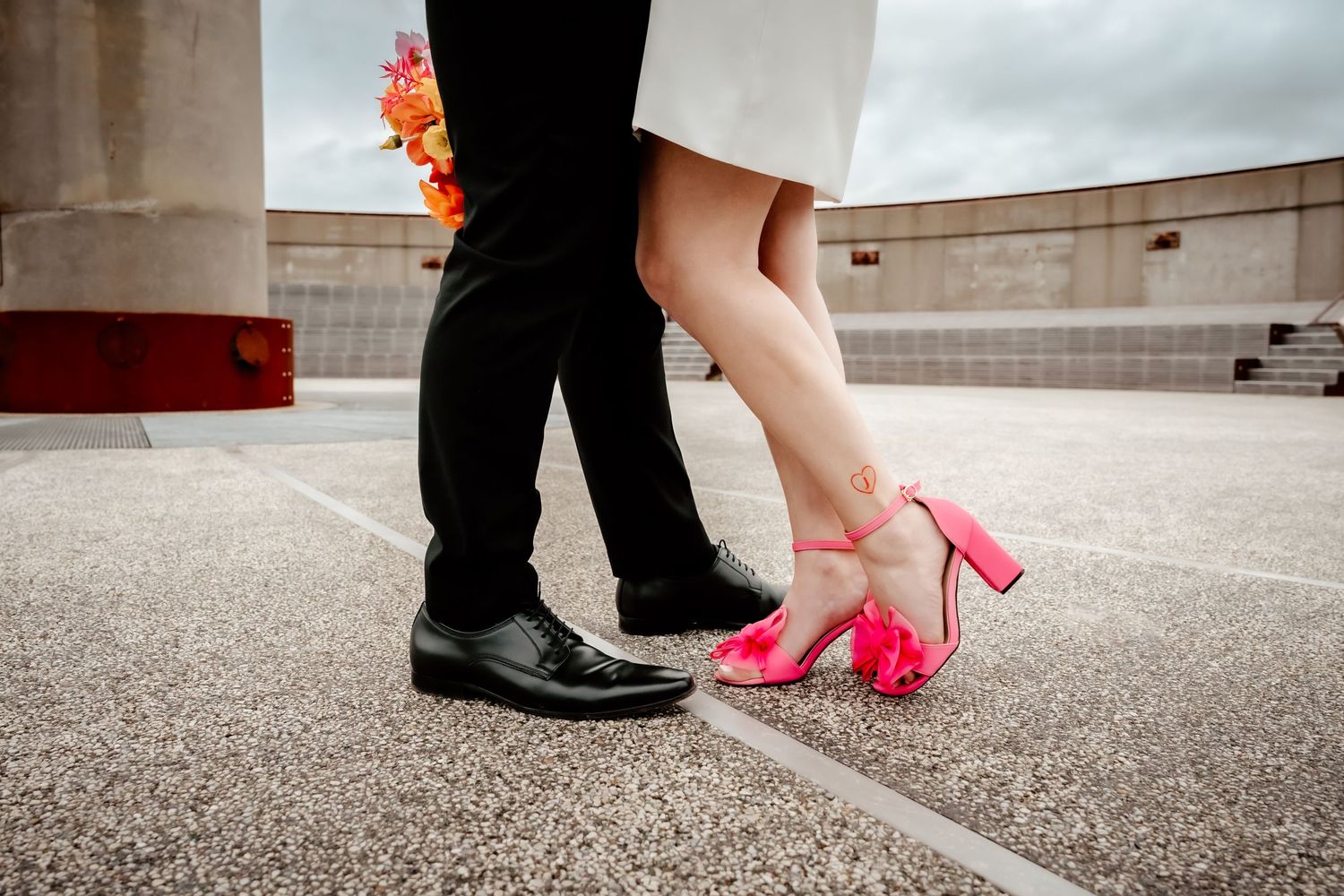 Wedding shoes detail showing red heels against black formal shoes on concrete.