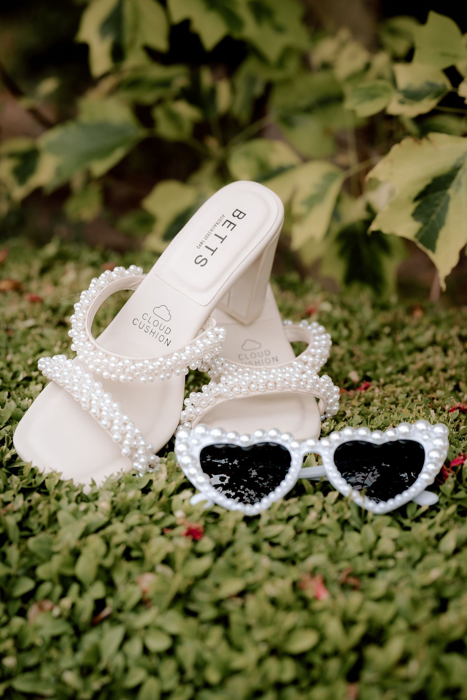 Pearl-embellished white wedding sandals and heart-shaped sunglasses arranged on green foliage.
