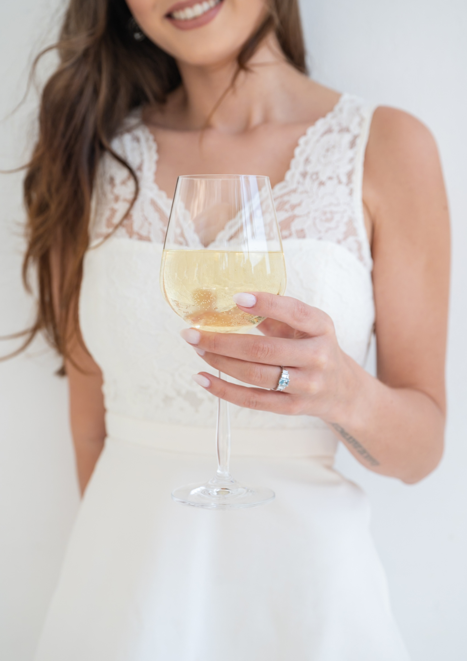 Person in white lace dress holding champagne glass against white background in elegant celebration setting.