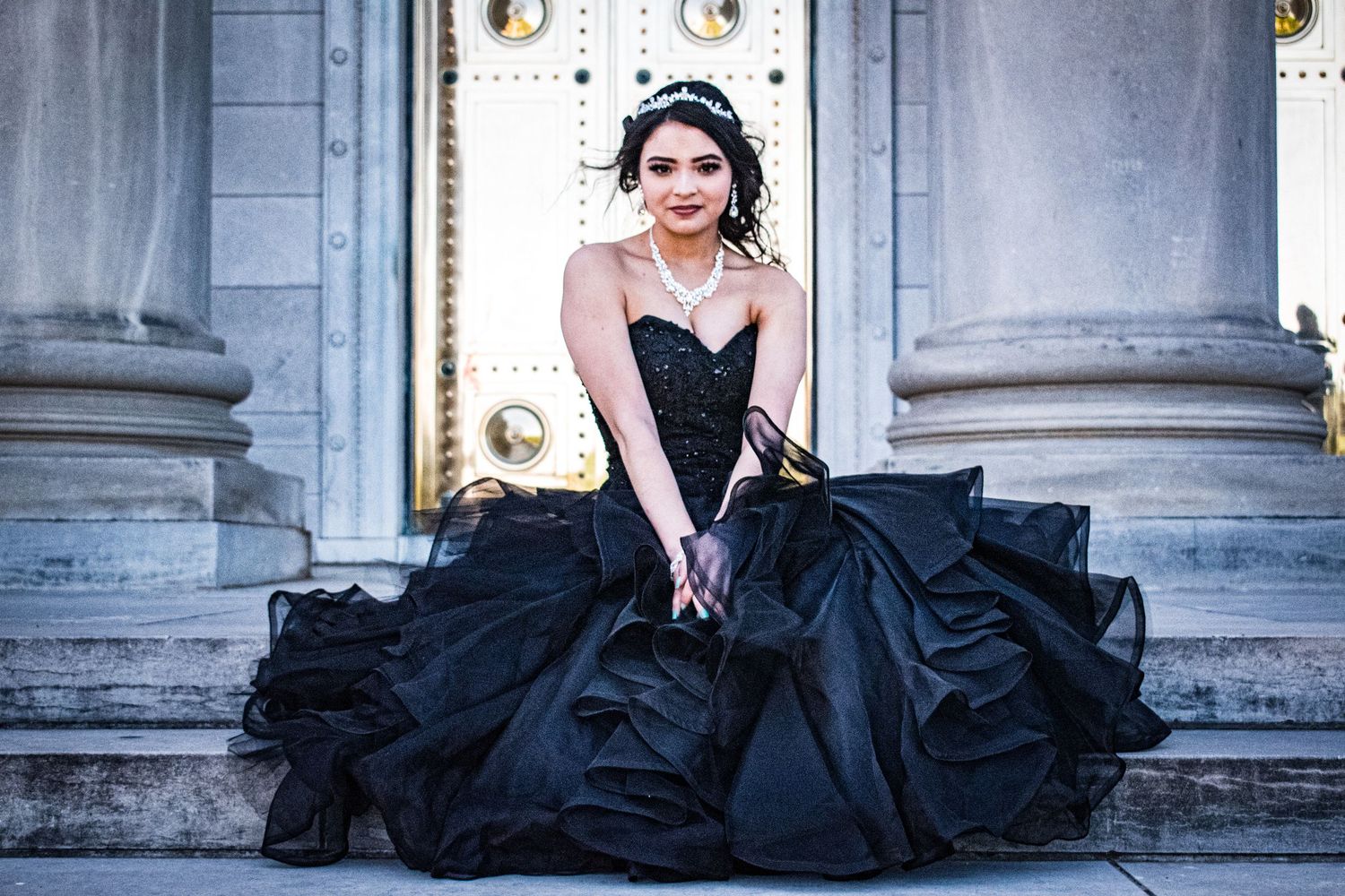 Young woman in elegant black ball gown seated on steps of grand building with columns.