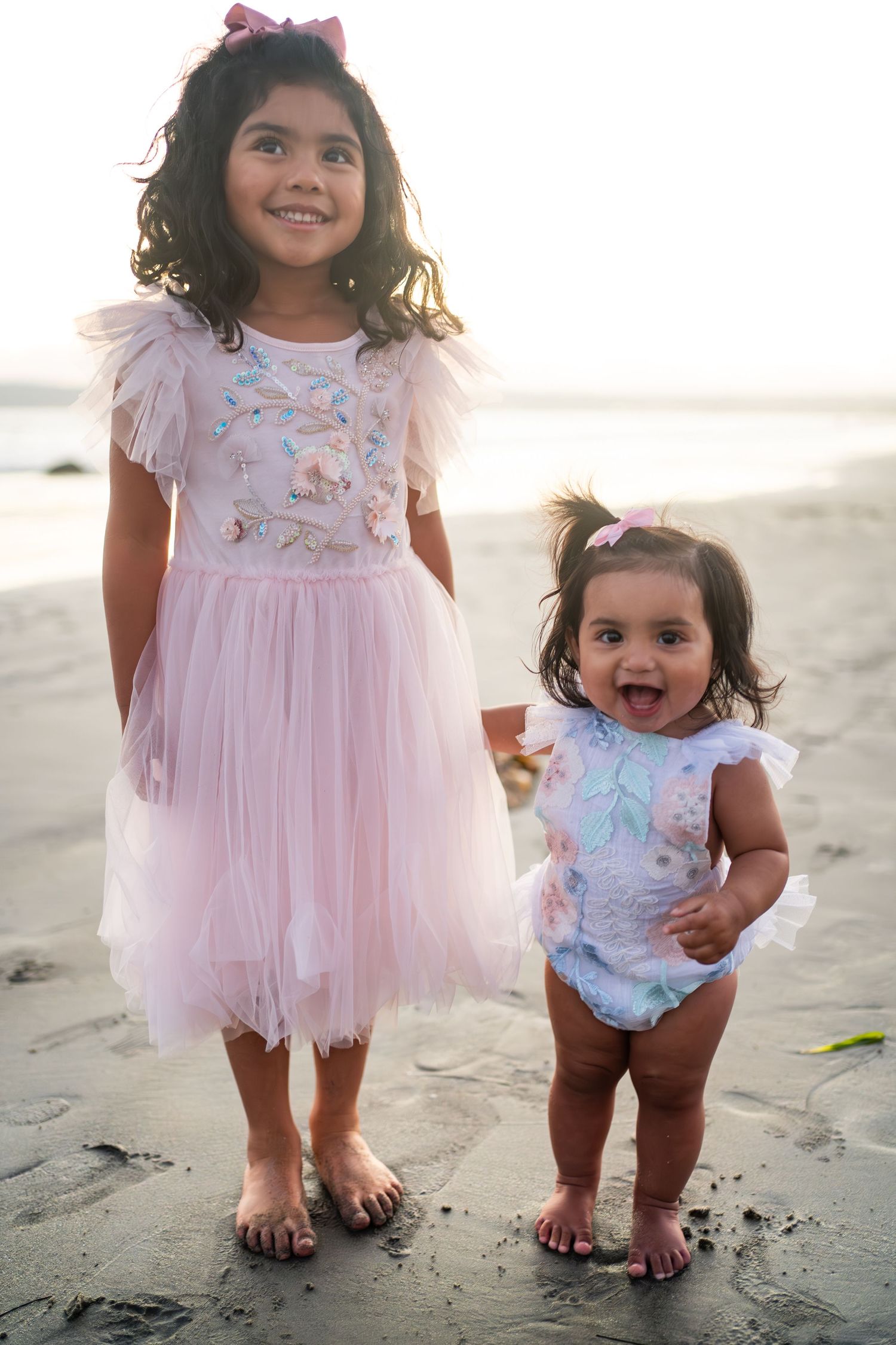 Two children in pink floral outfits stand barefoot on a sunny beach, one wearing a tulle dress and one in a romper.