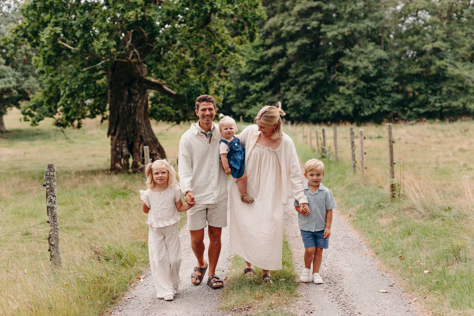 Children in light colored clothing run and play together on a tree-lined dirt path in a natural outdoor setting.