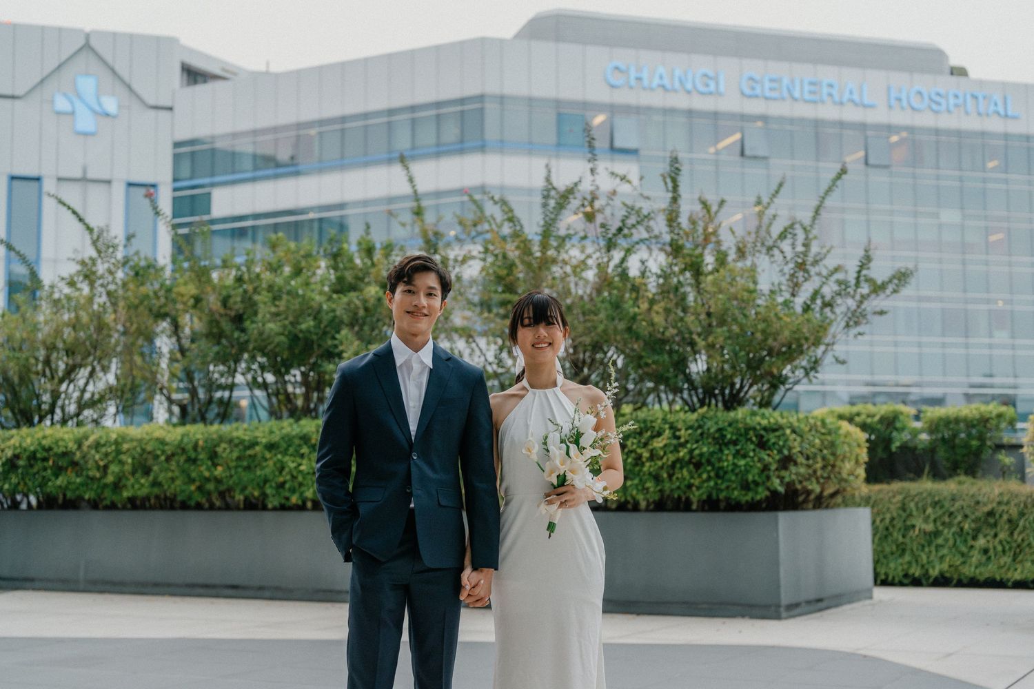 Wedding couple poses in front of a modern glass office building with landscaped grounds.
