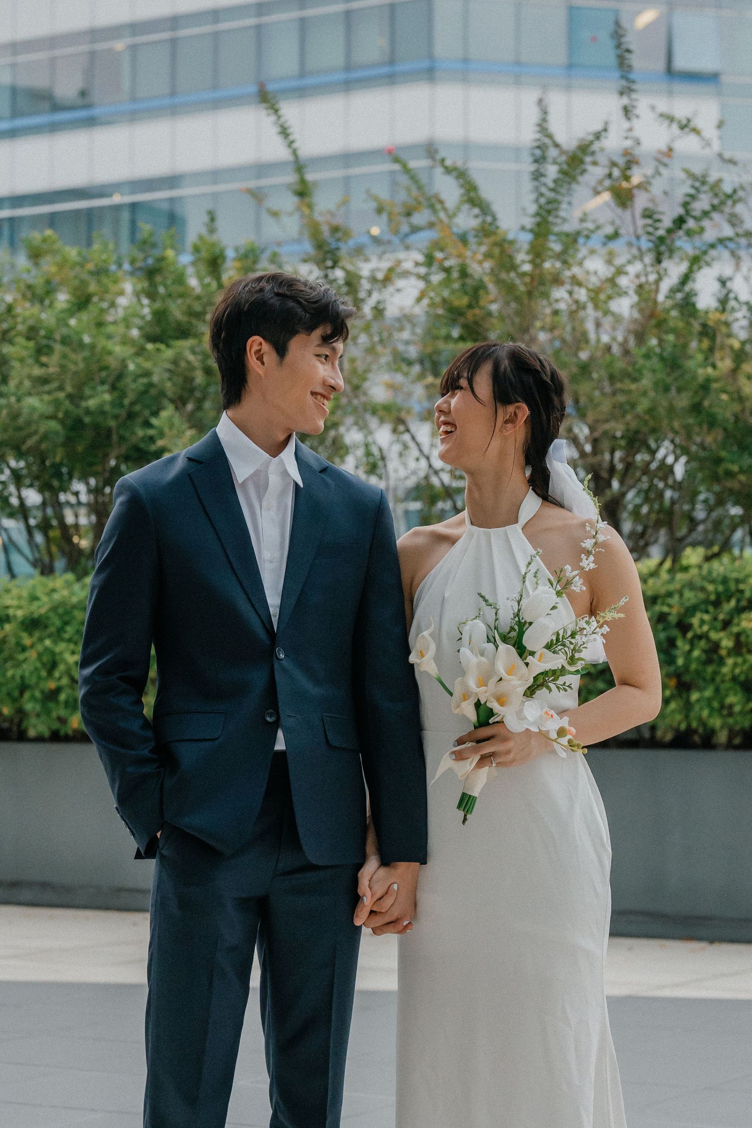 Elegant couple in wedding attire smiling at each other against modern architecture.