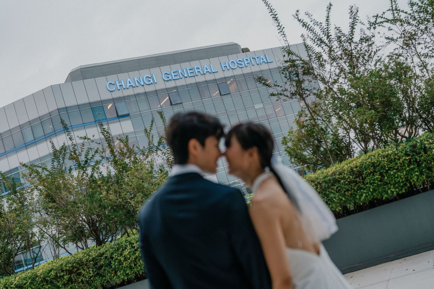 A gentle wedding moment captured in front of a modern hospital building with landscaping.
