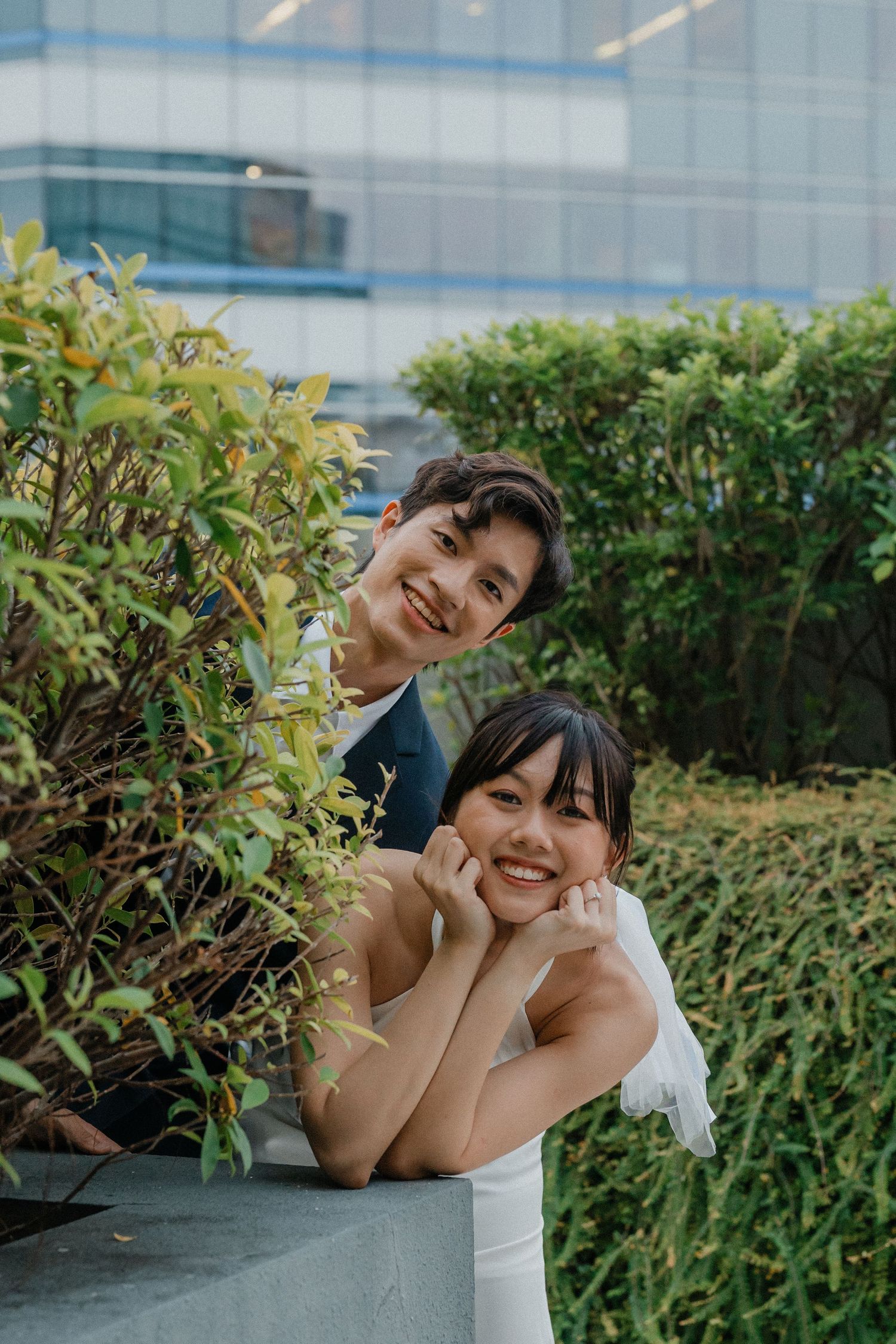 A cheerful wedding photo with the couple posing by garden shrubs near a hospital building.