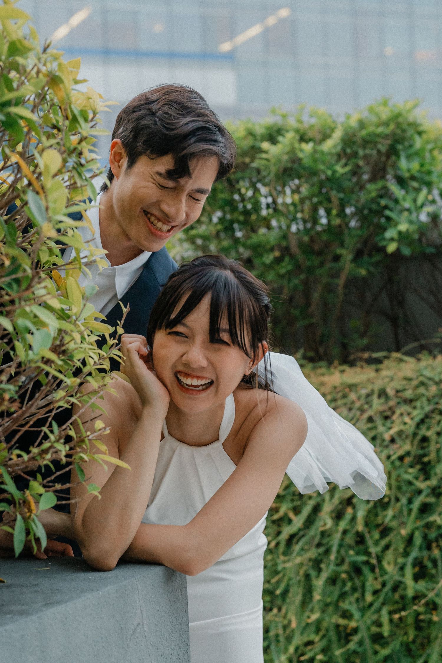 A sweet wedding moment captured by green shrubs with a modern building backdrop.