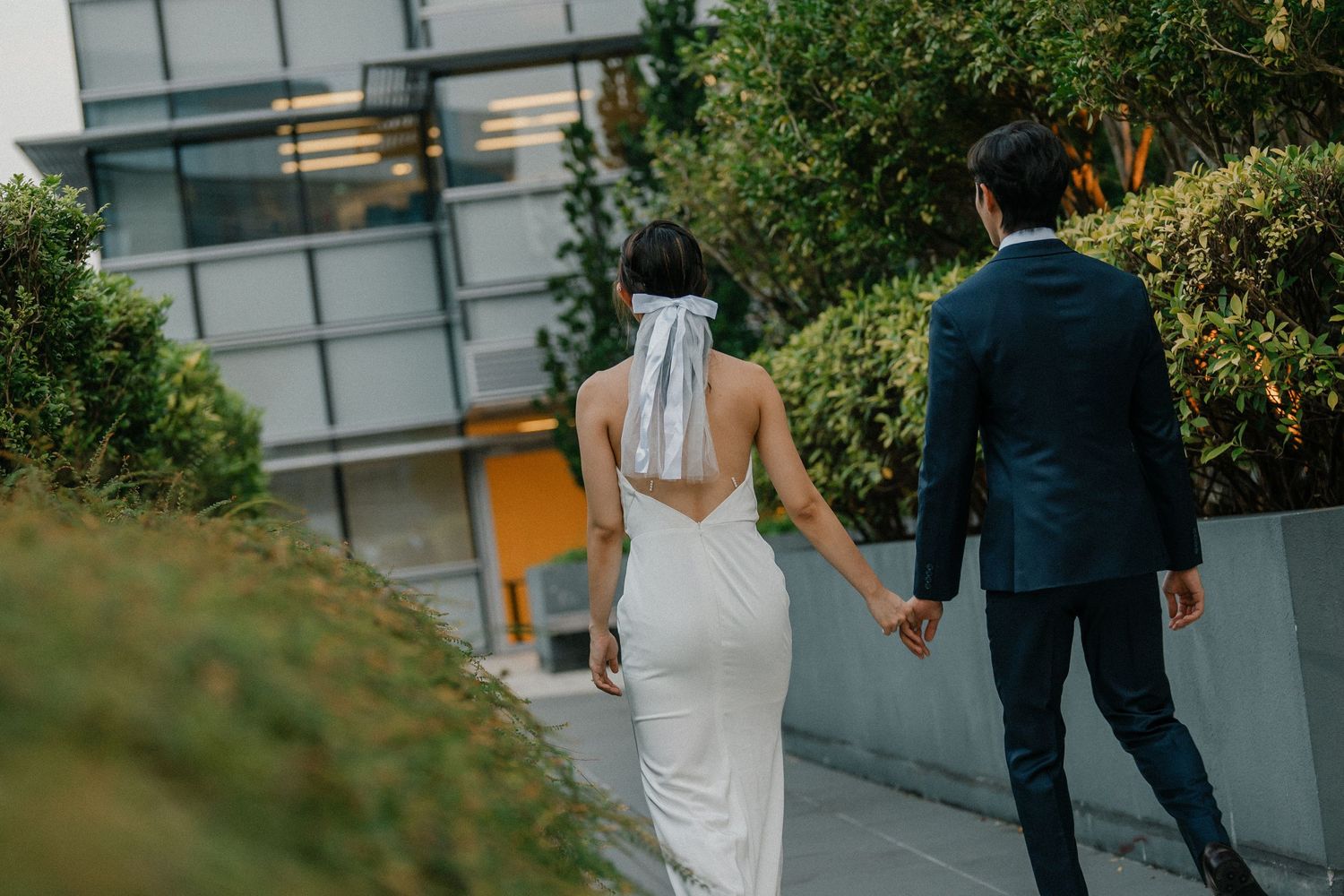 A wedding couple walks hand in hand along a garden path by a modern building.