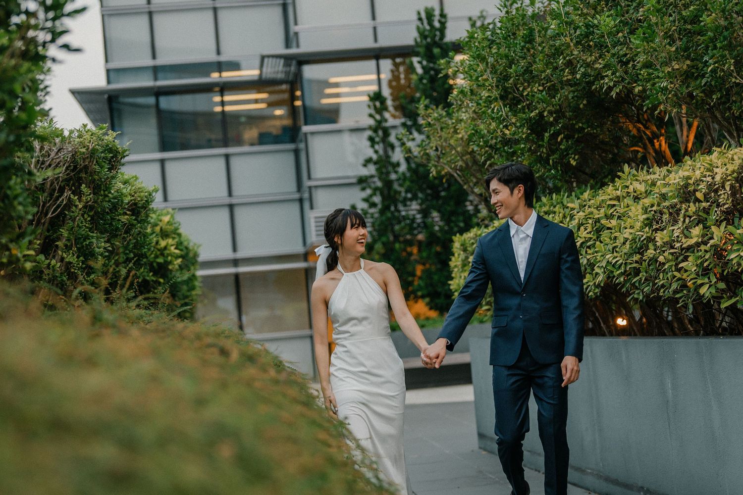 A couple in wedding attire walk hand in hand along a pathway next to a modern glass building with greenery.
