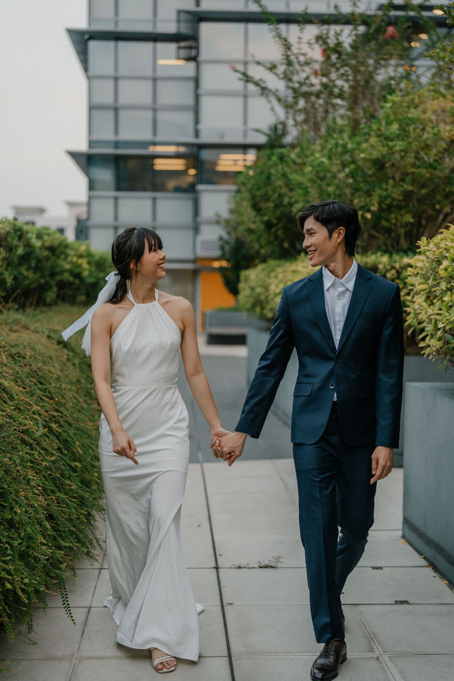 A couple in formal attire walks together on a concrete path beside a modern building with landscaping.