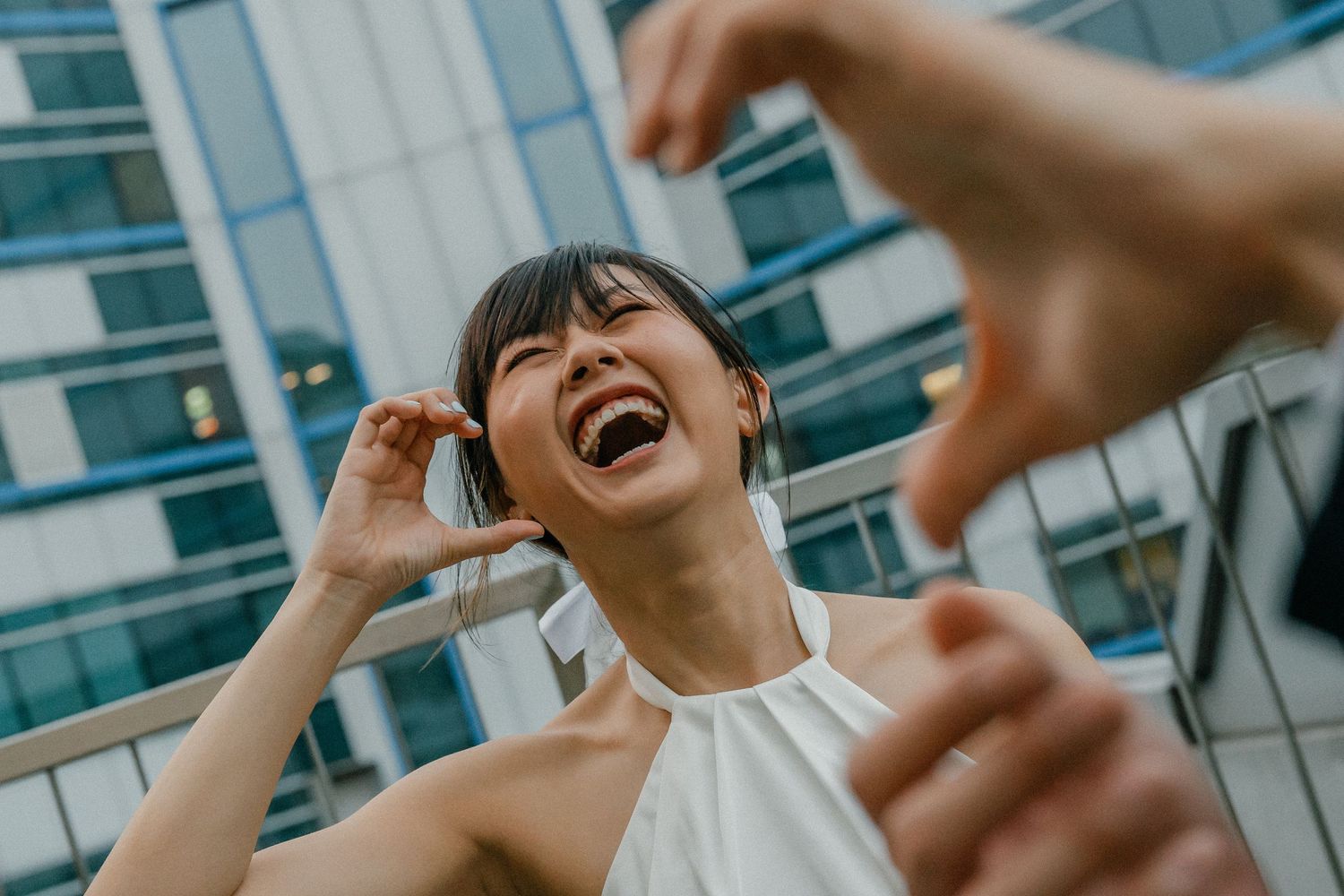 A joyful person in white dress laughs while making hand gestures in front of an urban building.