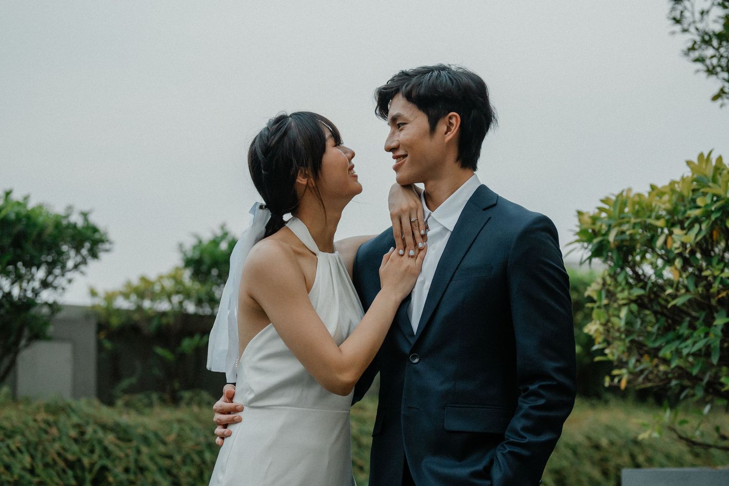 A couple in wedding attire embraces tenderly against a backdrop of green landscaping.