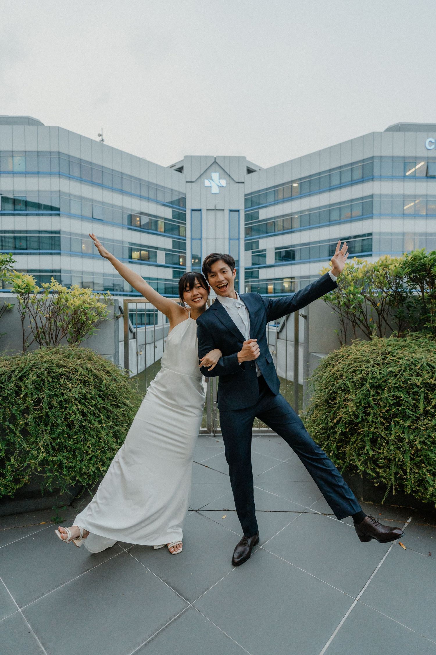 Newlyweds strike a fun pose with arms outstretched in front of a modern office building.