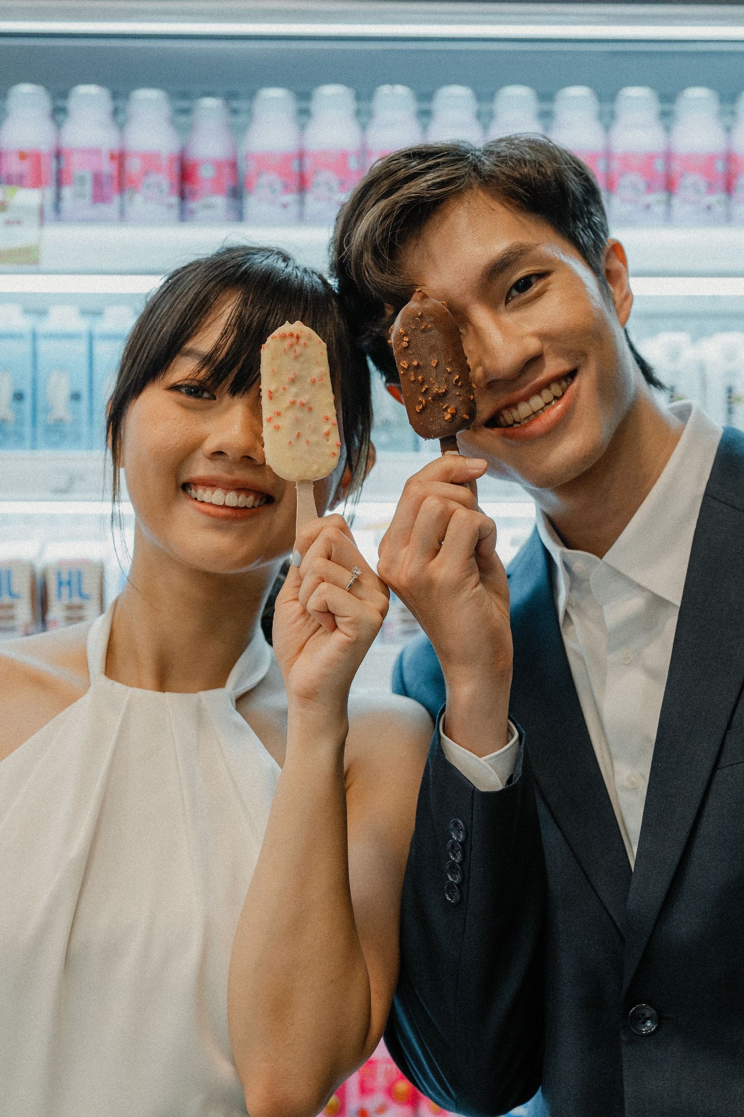 Couple poses playfully with ice cream bars in front of a refrigerator display.