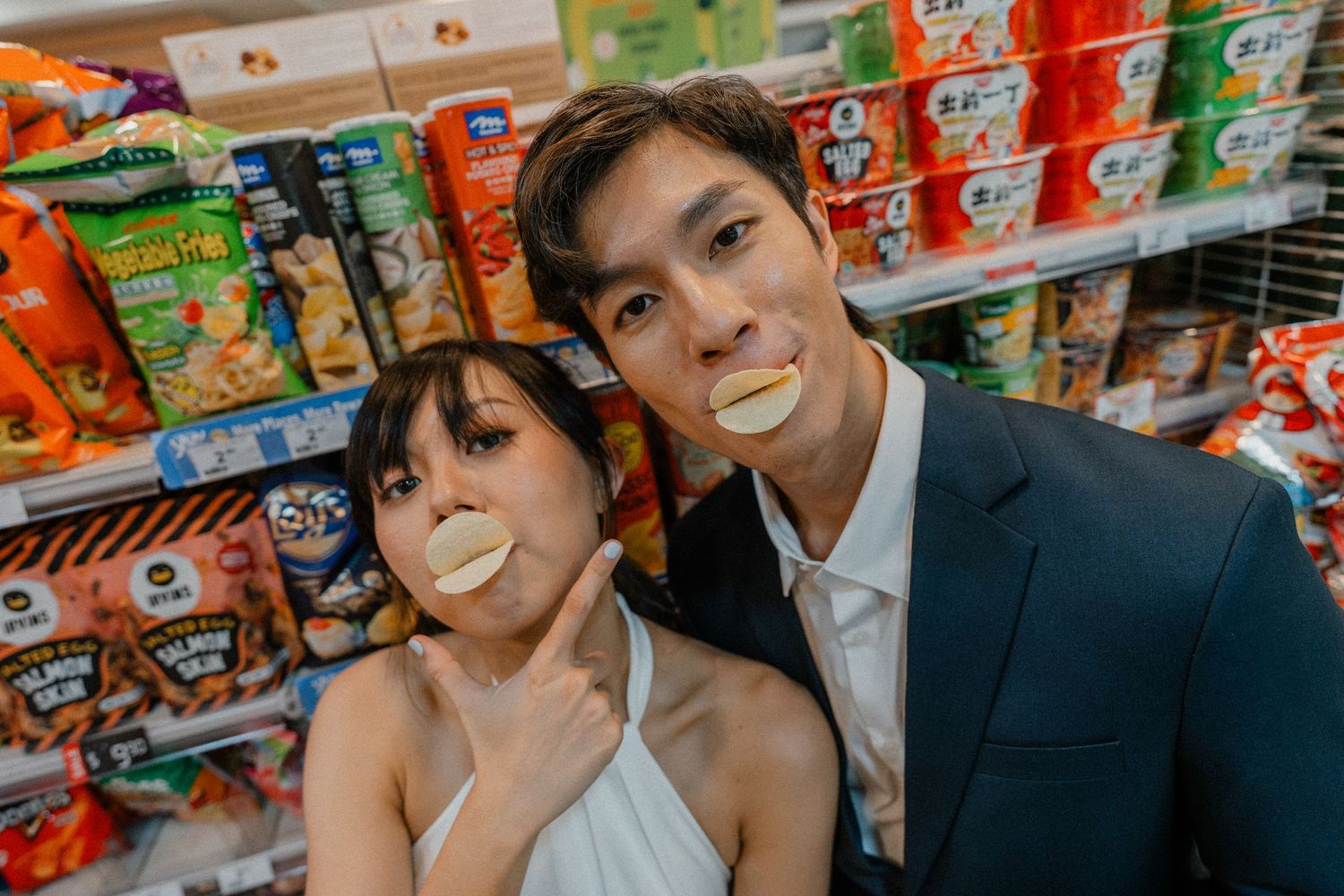 Couple poses playfully with potato chips in their mouths at a convenience store snack aisle.