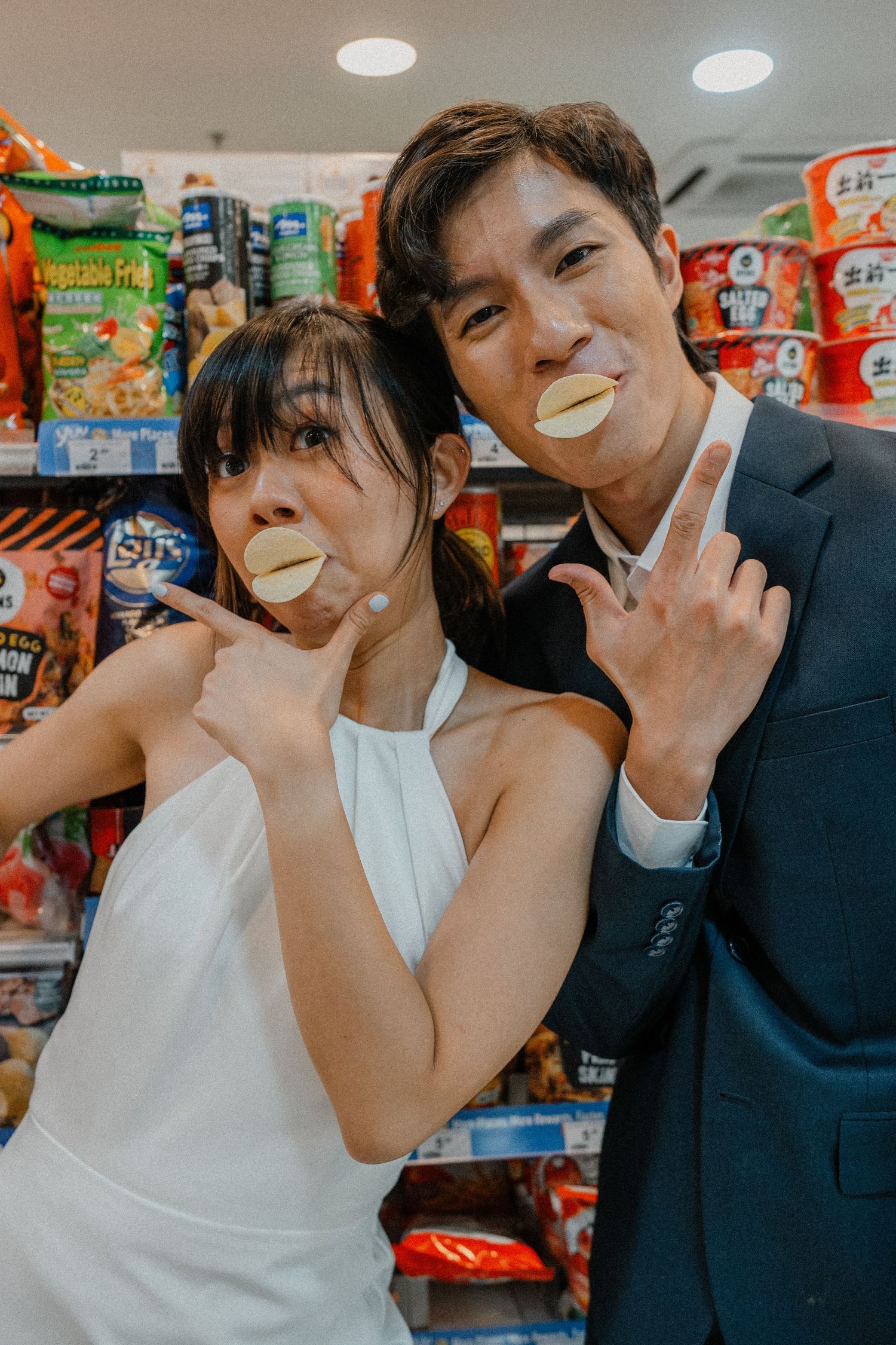 Playful couple making peace signs with snacks in their mouths at a grocery store.