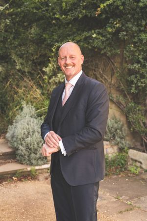 A gentleman in a dark suit poses outdoors in a garden setting.