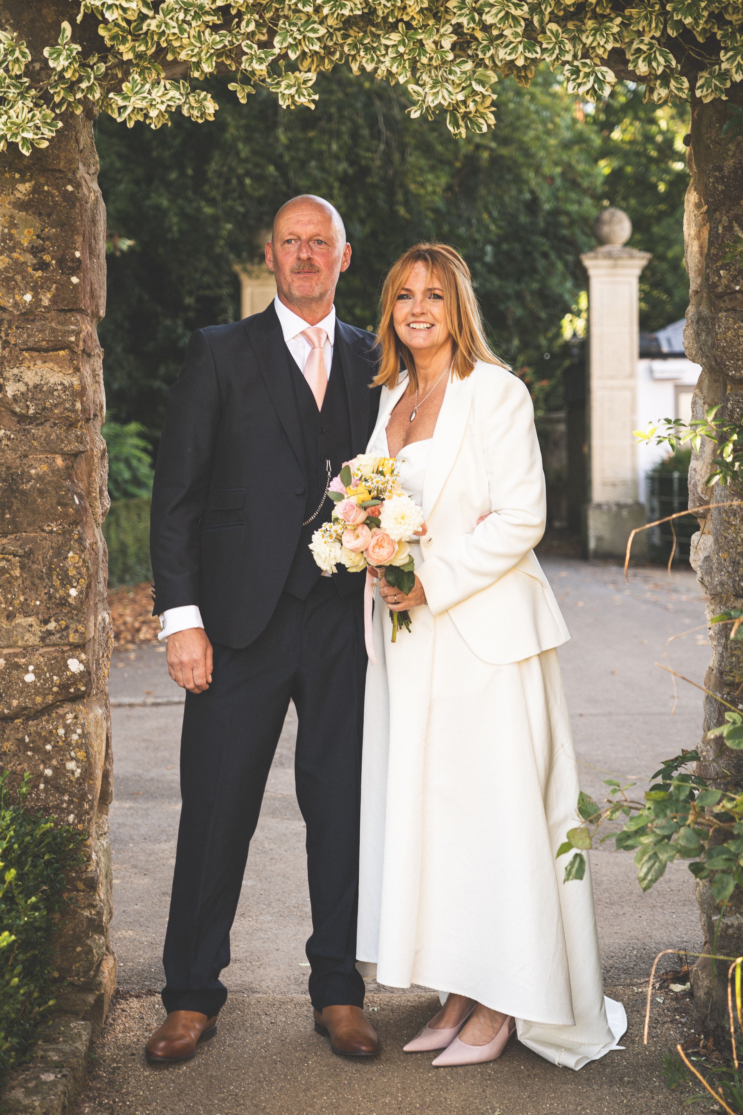 Couple in formal wedding attire walking along tree-lined pathway.