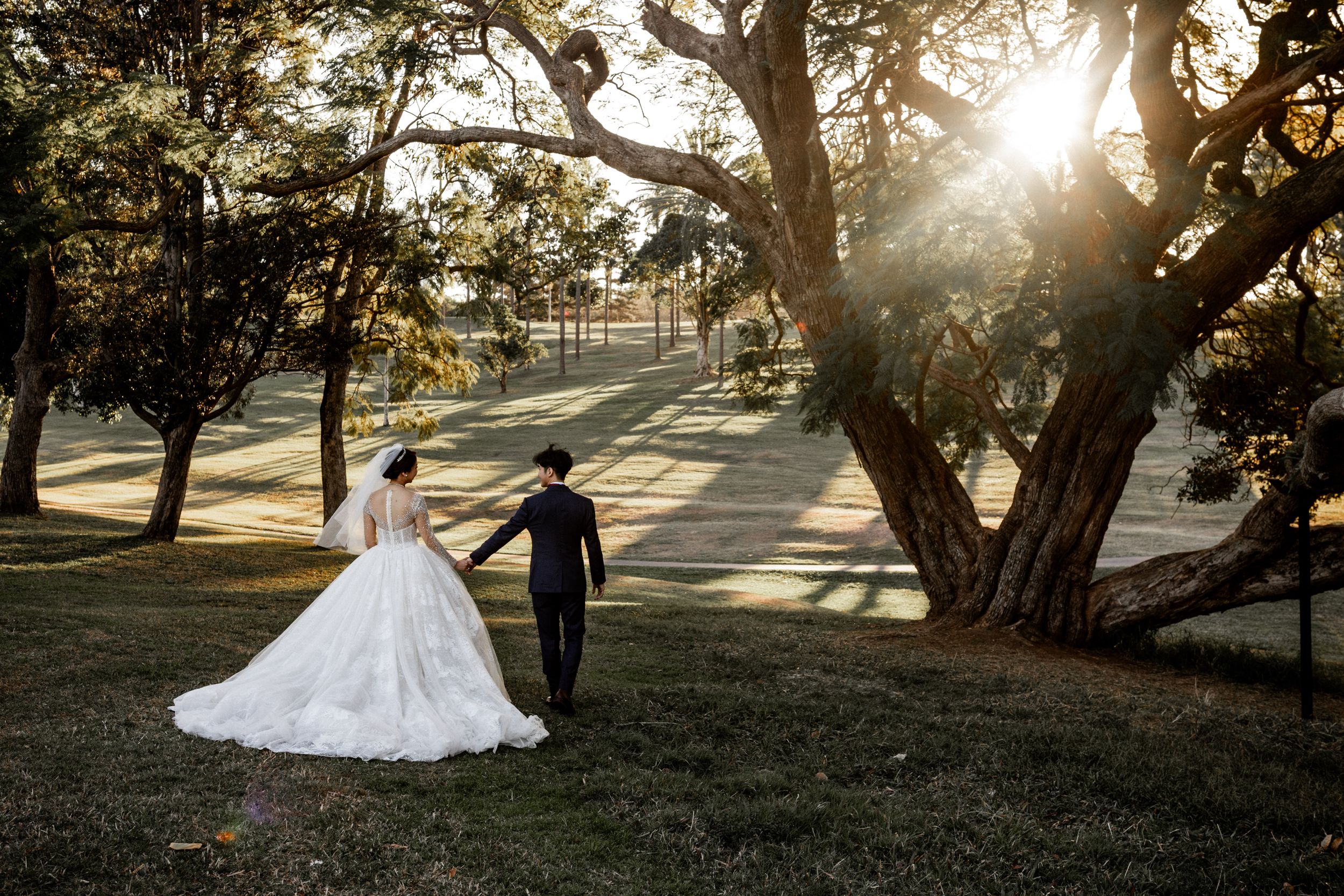 Newlyweds walk hand in hand through park during golden sunset beneath large trees.