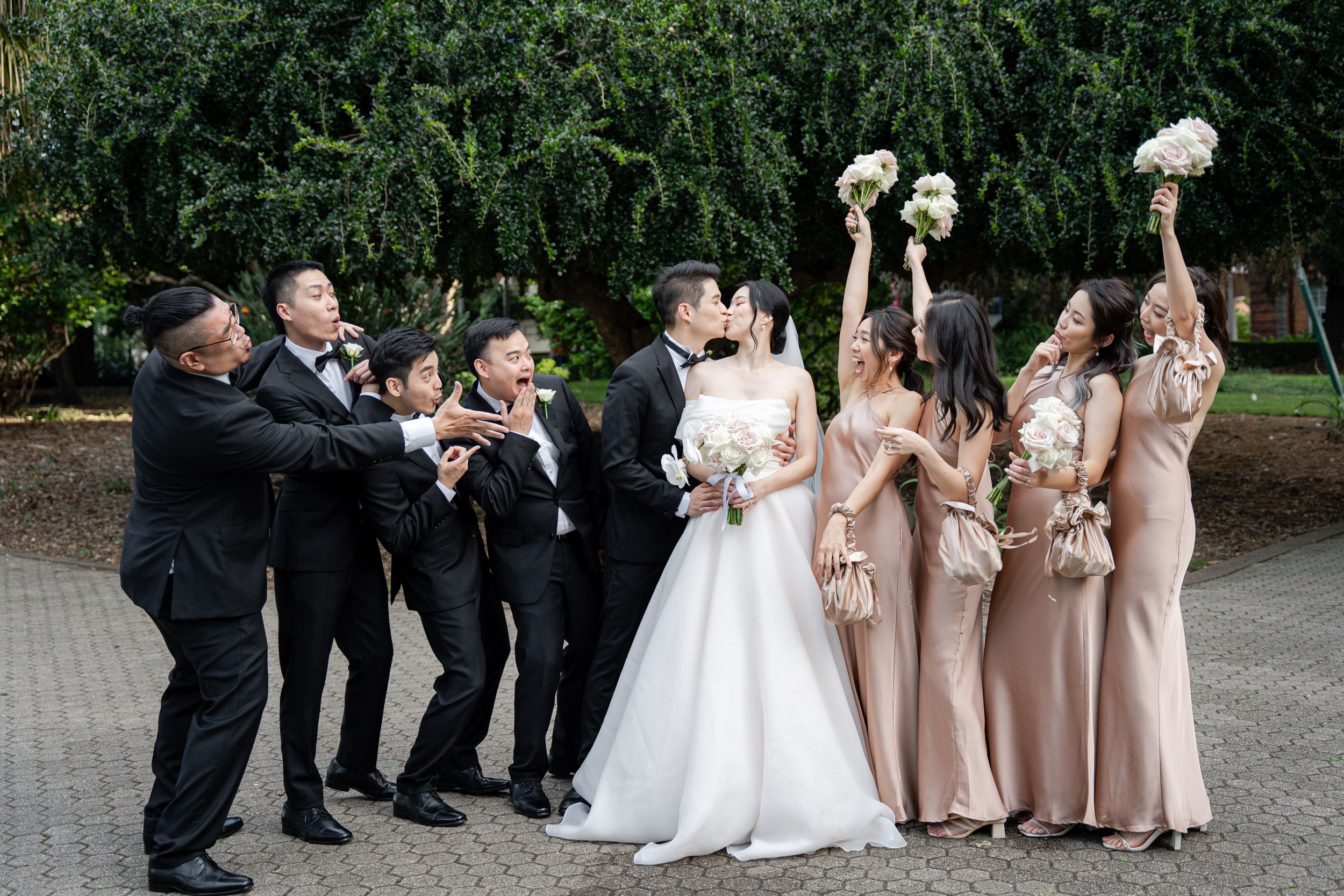 Wedding party strikes fun pose with bridesmaids raising bouquets while groomsmen lean to side.
