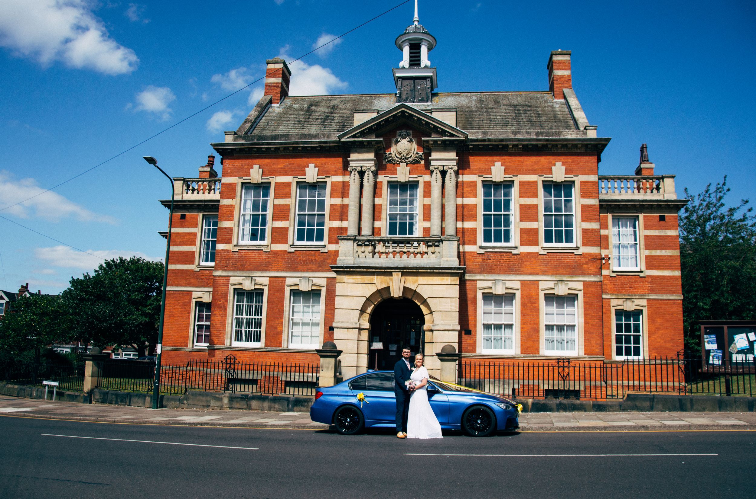Wedding couple outside historic red brick building under blue sky.