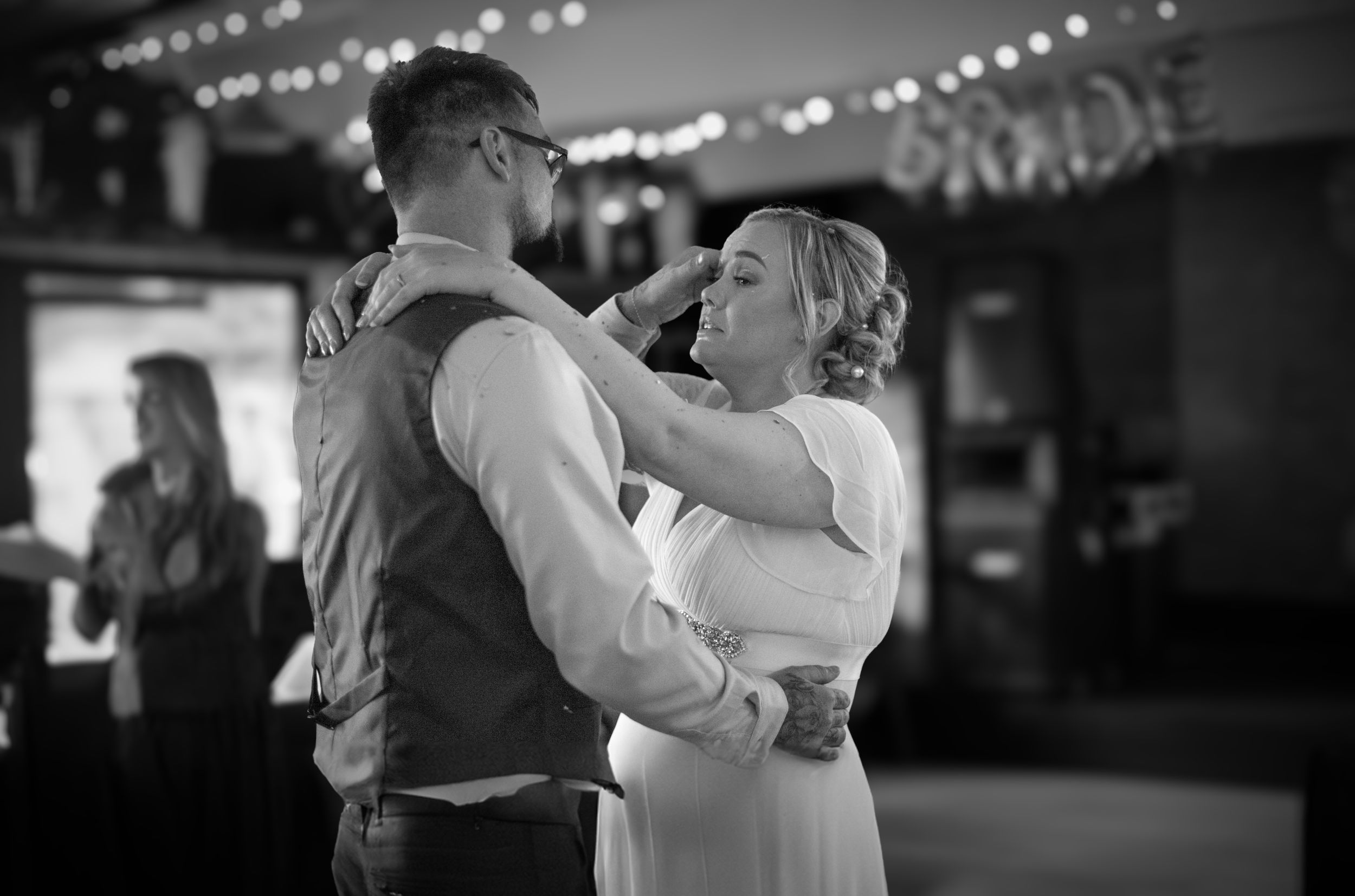 A romantic black and white photo of a couple slow dancing under string lights at their wedding reception.