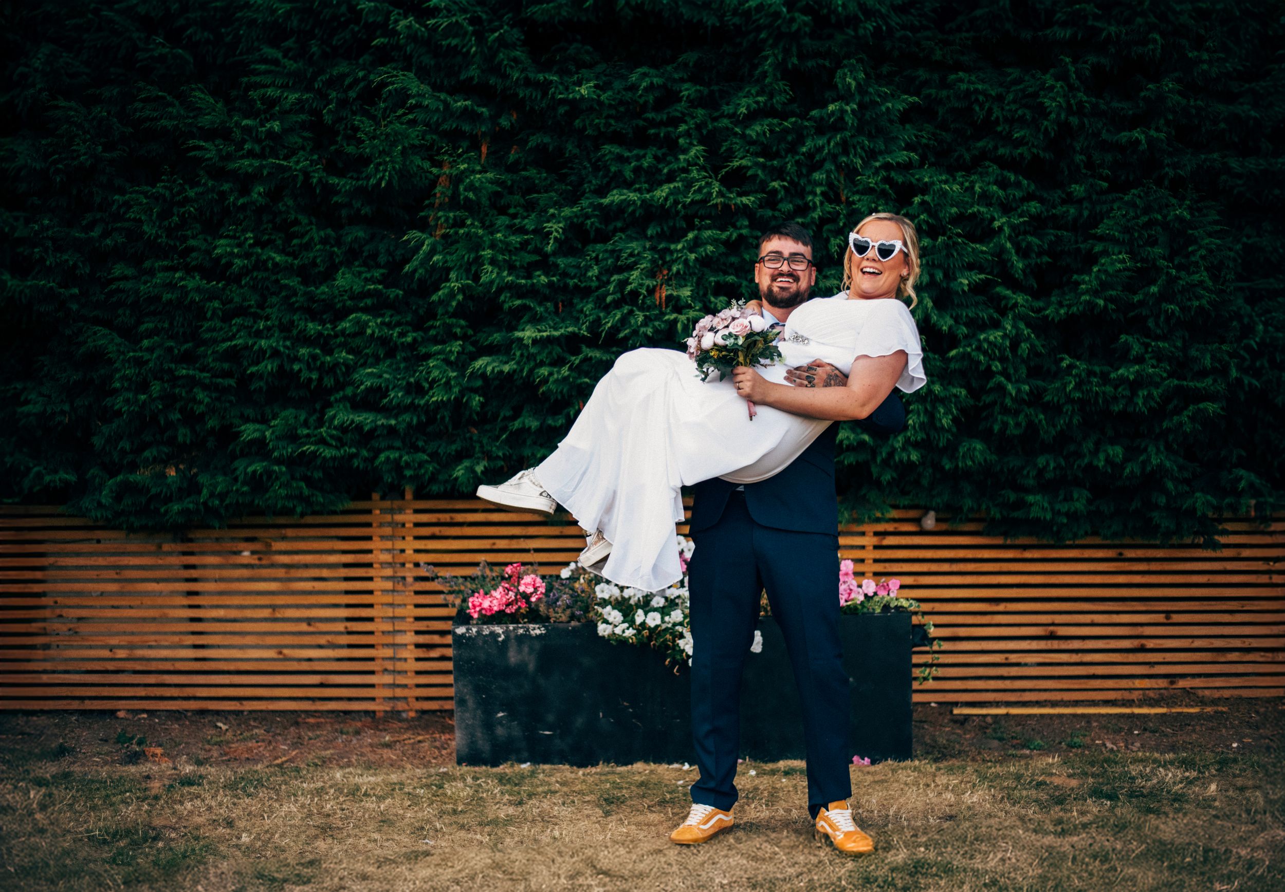 A couple in wedding attire poses playfully in front of a wooden fence and greenery backdrop.