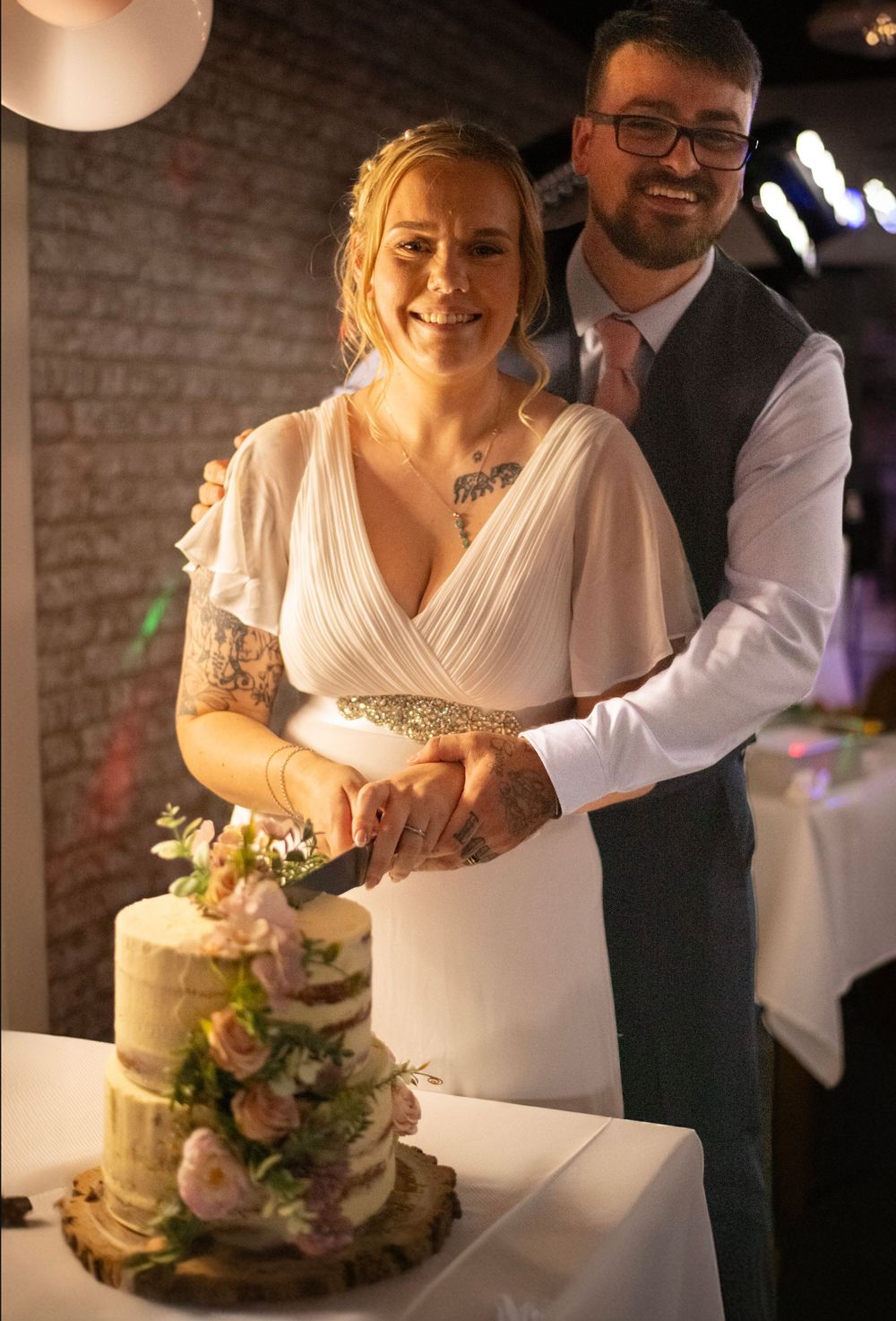 A couple cutting their elegant three-tiered wedding cake decorated with fresh flowers in a brick-walled venue.