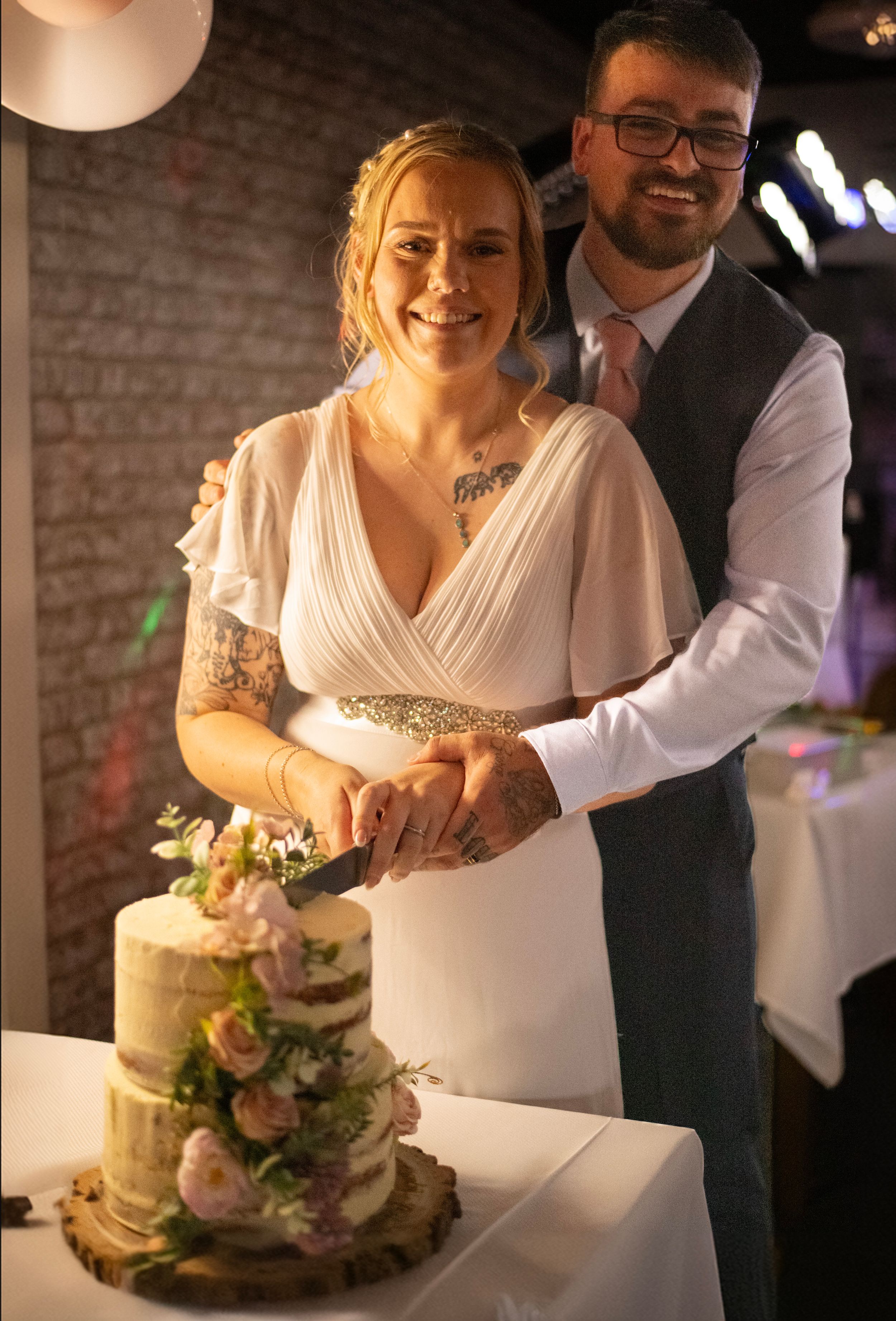 A couple cutting their elegant three-tiered wedding cake decorated with fresh flowers in a brick-walled venue.
