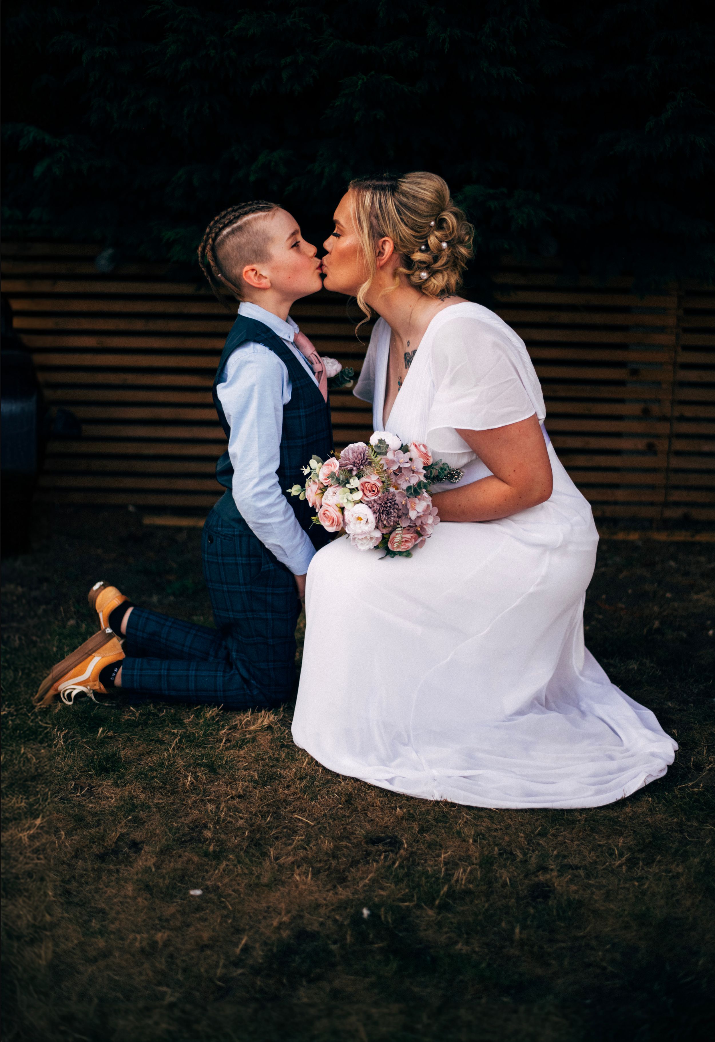 A romantic moment captured between a couple in wedding attire kneeling on grass with a bouquet.