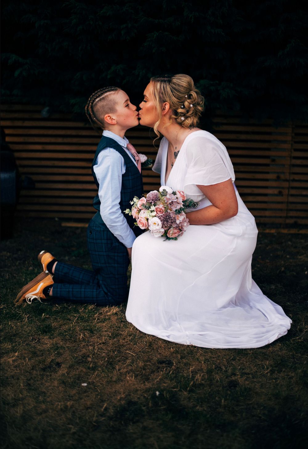 A romantic moment captured between a couple in wedding attire kneeling on grass with a bouquet.