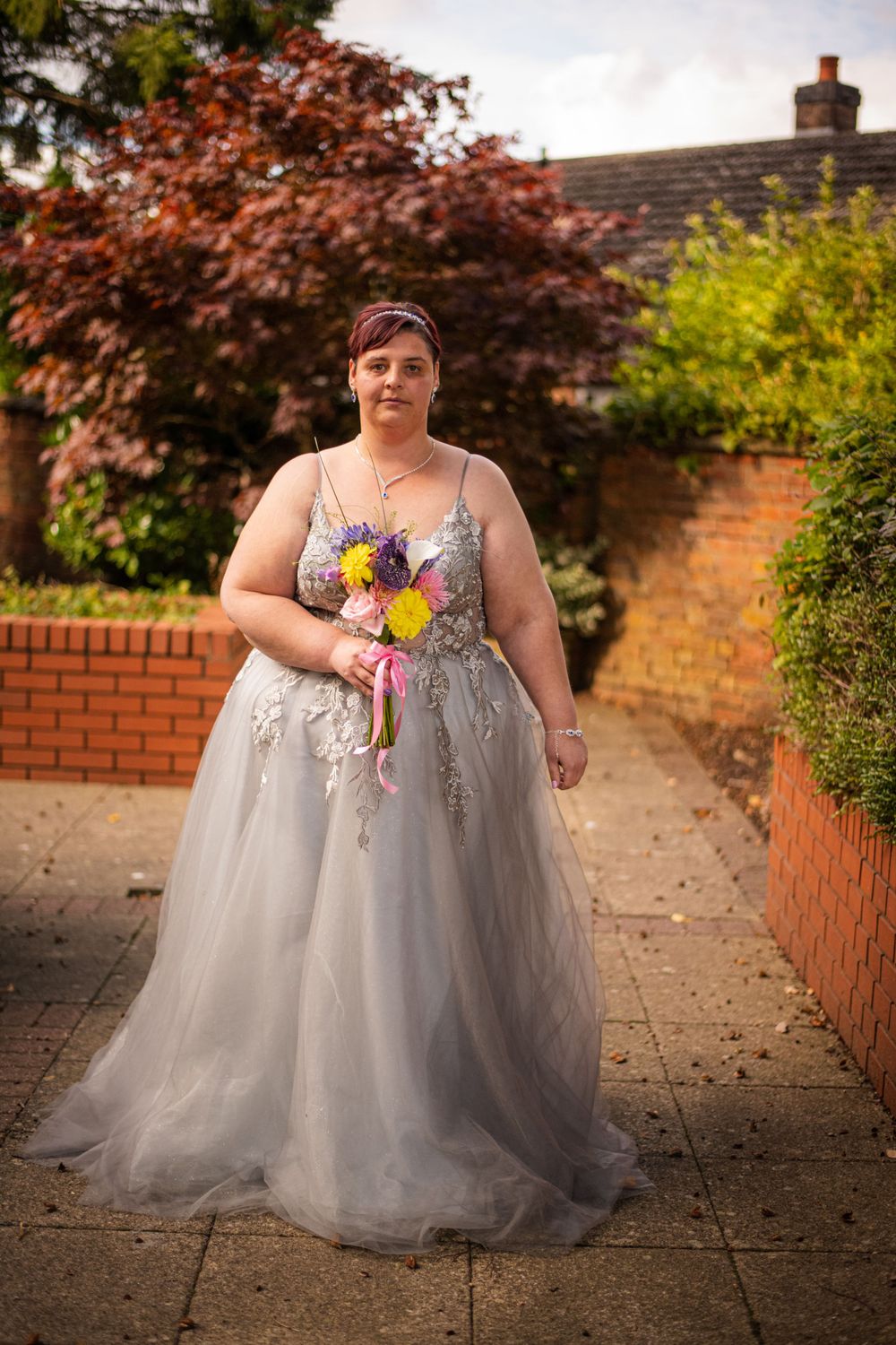 A person in a gray tulle wedding gown holding colorful flowers poses in a garden setting.