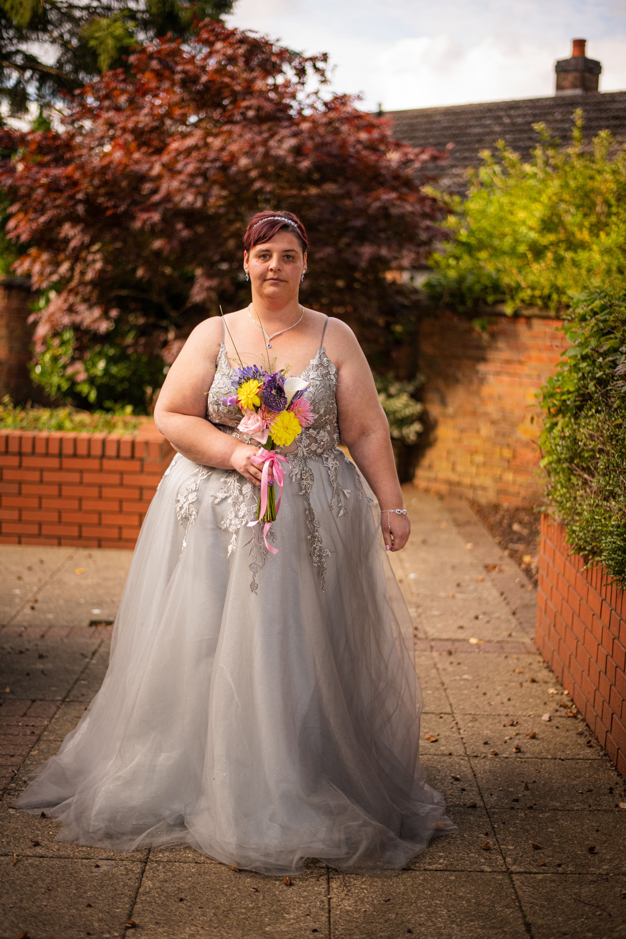A person in a gray tulle wedding gown holding colorful flowers poses in a garden setting.