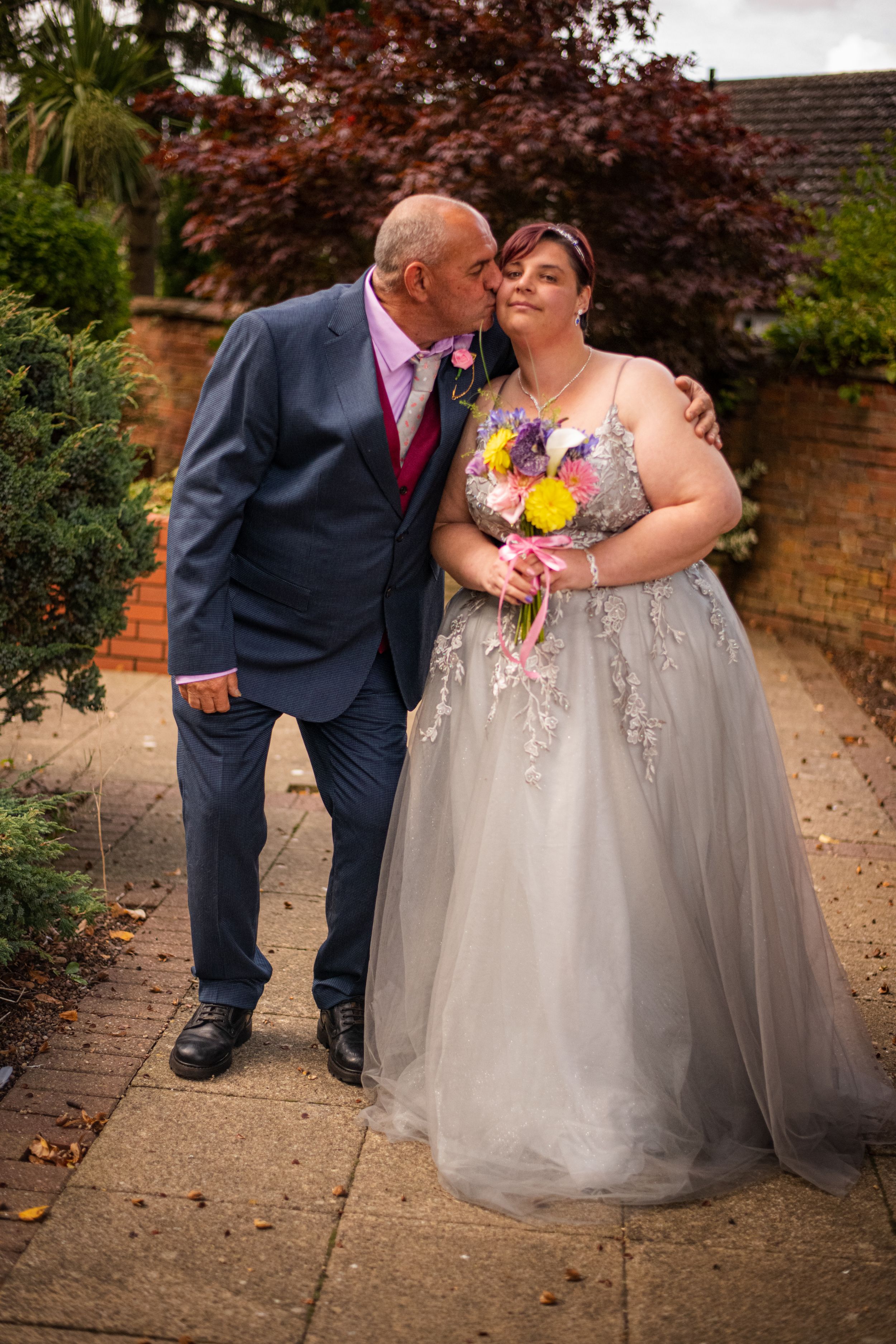 A couple shares a tender moment in formal wedding attire in front of a brick garden wall.