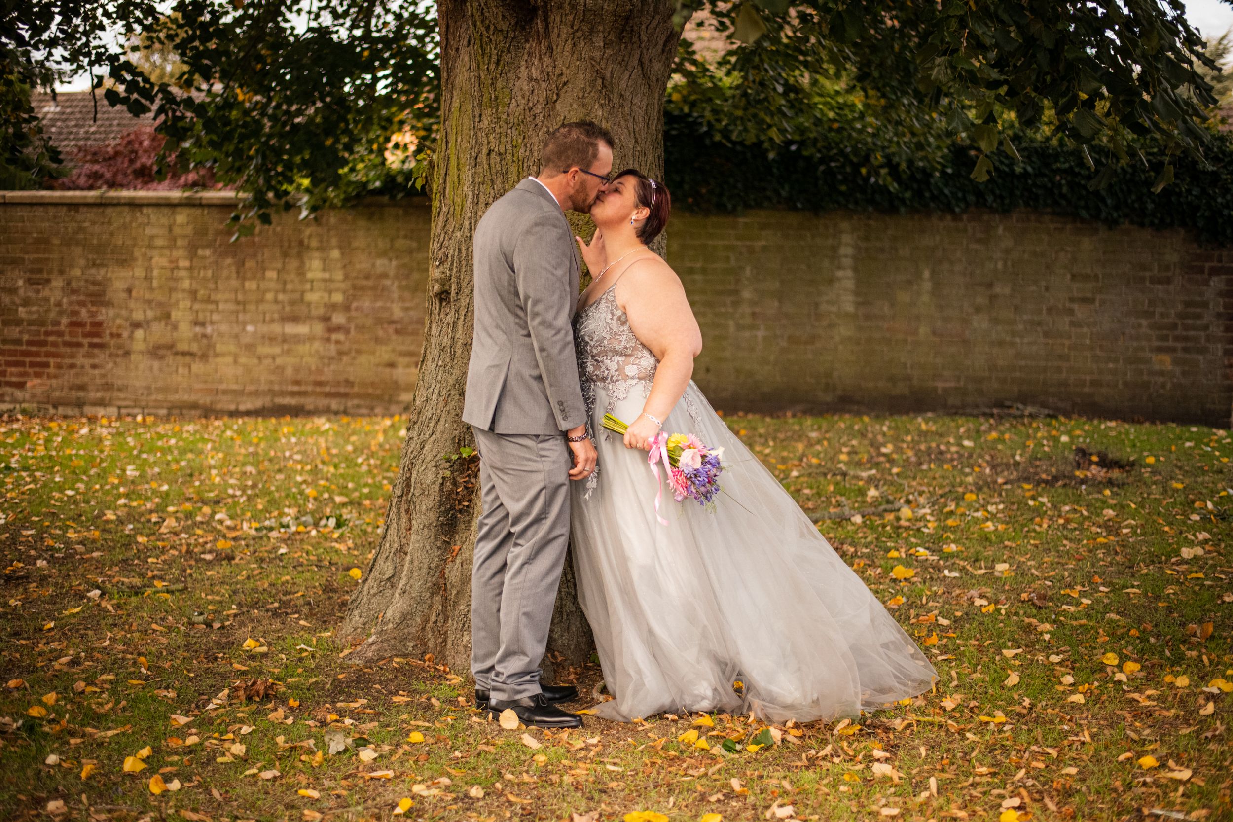 A romantic wedding photo of a couple leaning against a tree with fallen autumn leaves.