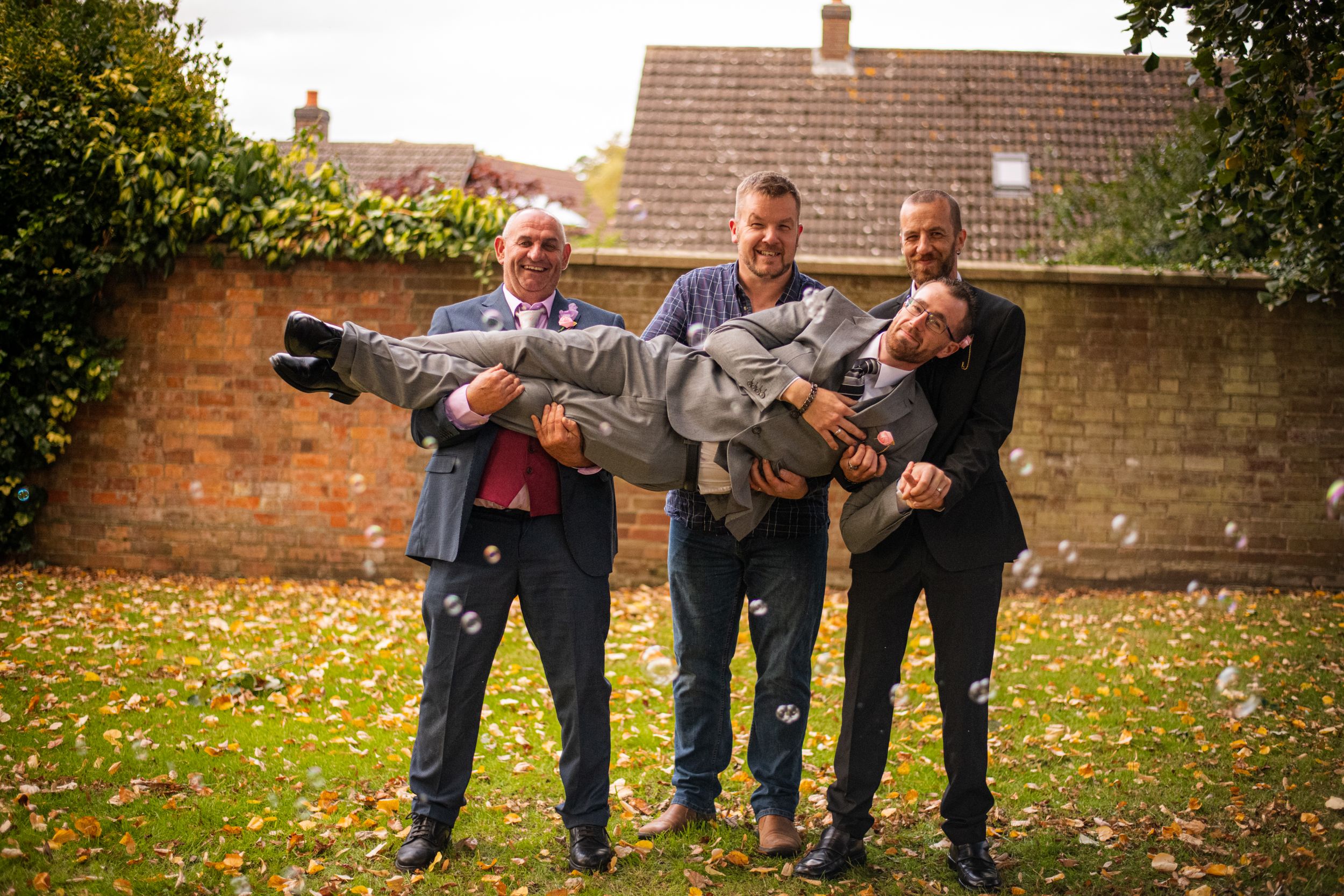 Three people playfully carrying someone in formal attire in a garden setting.
