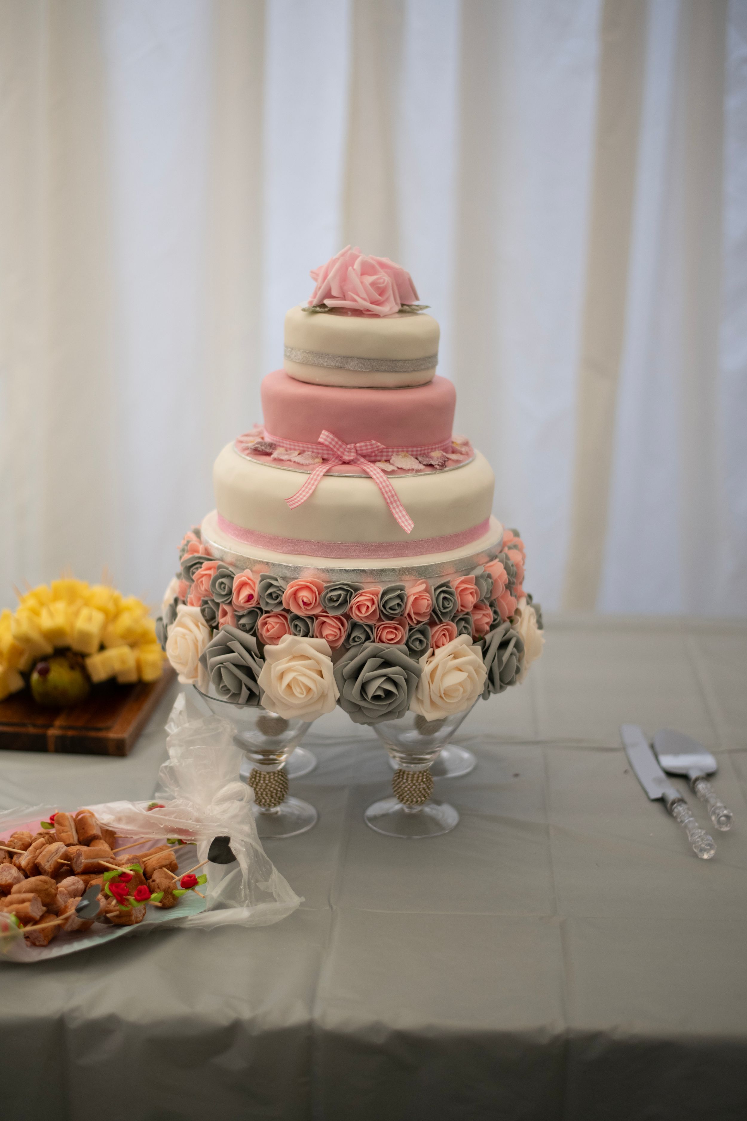 Elegant three-tier wedding cake with pink and gray roses displayed on a crystal cake stand with refreshments nearby.